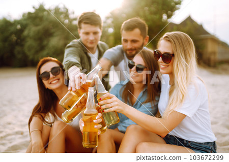 Group of friends cheers and drink beers on the beach. Young friends relaxing and having picnic. Suummertime. 103672299