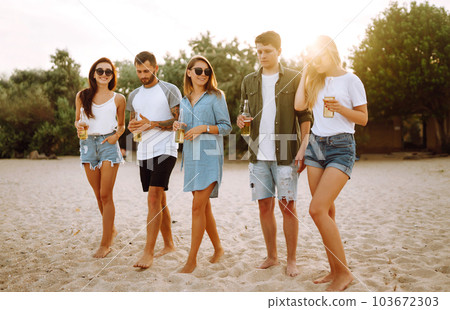 Group of friends cheers and drink beers on the beach. Young friends relaxing and having picnic. Suummertime. 103672303