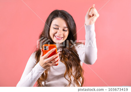 Long-haired lady with amused expression reads message on winning lottery on phone. Woman looks at won amount of money with shocking expression Long-haired lady with amused expression reads message on winning lottery on phone. Woman looks at won amount of money with shocking expression 103673419