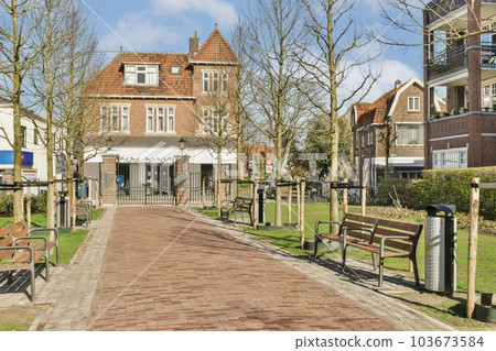 a street with benches and trees in the middle of the road, surrounded by buildings on both sides there is a blue sky above 103673584