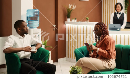 Happy newlywed couple on their honeymoon trip waiting in hotel lobby to be checked in while helpful bellhop employee carries luggage to their room. Husband and wife enjoying coffee in resort foyer 103674731