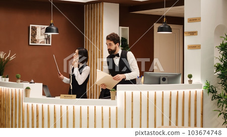 Teamworking professional receptionists checking guests vacation packages at check in reception desk. Hotel personnel booking incoming tourists, working in stylish resort lounge 103674748
