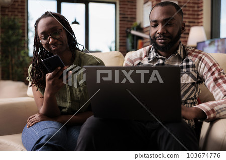 African american woman browsing social media on smartphone while boyfriend is working on laptop sitting on couch. Couple spending free time together using digital devices in home living room. African american woman browsing social media on smartphone while boyfriend is working on laptop sitting on couch. Couple spending free time together using digital devices in home living room. 103674776