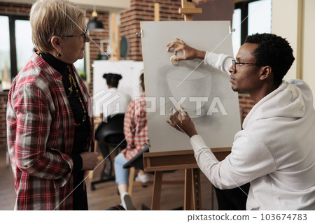 Senior woman taking up drawing class in art studio, listening to teacher. Young African American guy instructor sitting at easel demonstrating sketching technique. Learning new skill in retirement 103674783