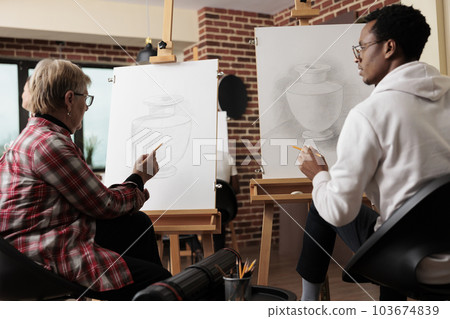 Diverse different generation people drawing on canvas and talking during creative art lesson in modern studio. Senior woman teaching African American student guy how to draw naturalistic still life Diverse different generation people drawing on canvas and talking during creative art lesson in modern studio. Senior woman teaching African American student guy how to draw naturalistic still life 103674839
