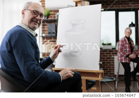 Happy senior man attending art workshop. Positive retired student sitting at easel and practicing drawing on canvas in class, enjoying to learn new things. Creative retirement concept 103674847