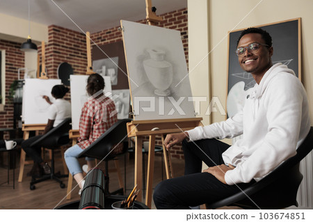 Portrait of happy young African American man student of art school sitting at easel smiling at camera, learning pencil drawing in class. Positive black guy enjoying creative hobby with friends 103674851