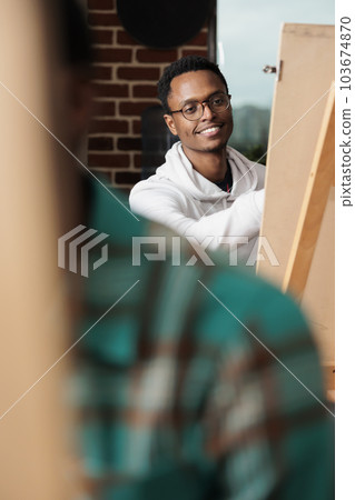 Smiling African American guy sits at easel enjoying creative activities in art studio. Happy young man visiting group painting session to learn artistic skills and meet people, drawing and having fun Smiling African American guy sits at easel enjoying creative activities in art studio. Happy young man visiting group painting session to learn artistic skills and meet people, drawing and having fun 103674870