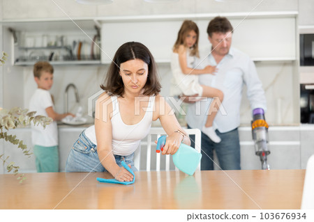 Wife cleans table with rag, husband and children clean kitchen in background 103676934