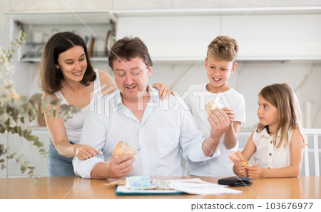 Adult man sitting at table distributes money to family 103676977