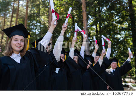 A group of graduates in robes raised their hands with diplomas outdoors. Elderly student. 103677759