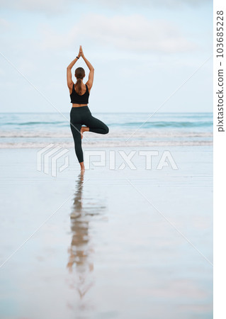 The steadiness in the tree pose helps you calm your mind. Rearview shot of a young woman doing a tree pose while practising yoga at the beach. 103685228