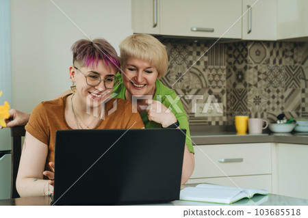 Adult modern mother and non-binary daughter using laptop together smiling Adult modern mother and non-binary daughter using laptop together smiling 103685518