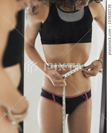 Anorexia, measuring tape and stomach of woman in mirror for weight loss, eating disorder and body image. Depression, mental health and sad with female at home for size obsession, diet and anxiety 103688536