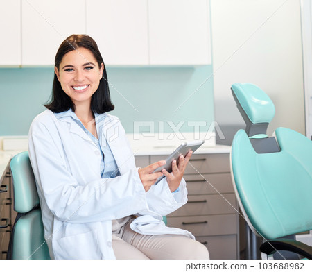 A beautiful, confident female dentist sitting in her doctors rooms. A confident, brunette woman working on a digital tablet while sitting in her office. Dental health resources are easy to find online 103688982