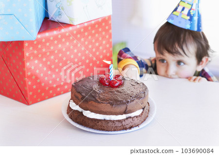 Birthday cake, kid and curious of food at party with gifts and dessert at home with a hungry child. Chocolate, sneaky and snack to celebrate a boy at a party feeling temptation for sweets in house 103690084