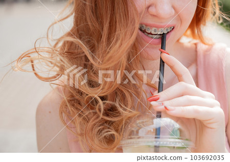 Young beautiful red-haired woman with braces drinks cooling lemonade outdoors in summer. Portrait of a smiling girl with freckles. 103692035
