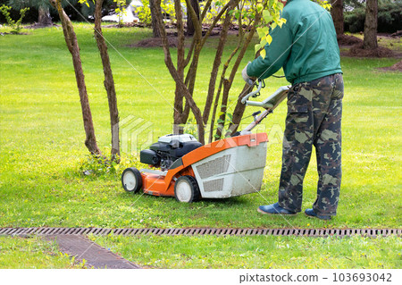 A gardener in camouflage clothing mows a grassy lawn with a petrol lawn mower on a spring day. A gardener in camouflage clothing mows a grassy lawn with a petrol lawn mower on a spring day. 103693042