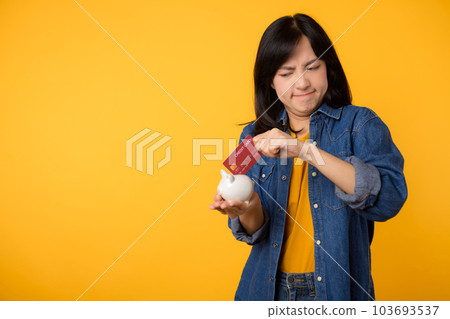 Unhappy asian young woman wearing yellow t-shirt denim shirt pulling credit card out of piggy bank isolated on yellow background. Payment debt financial concept. 103693537