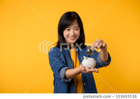 Happy young asian woman wearing yellow t-shirt denim shirt saving digital crypto currency to piggy bank isolated on yellow background. Digital money saving investment concept. 103693538