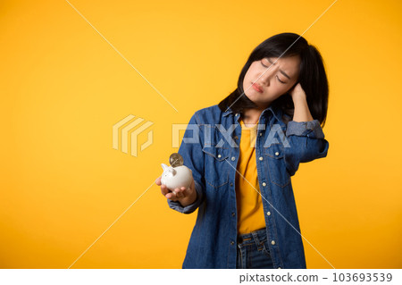 Unhappy asian young woman wearing yellow t-shirt denim shirt pulling digital coin crypto currency out of piggy bank isolated on yellow background. Payment digital money debt financial concept. 103693539