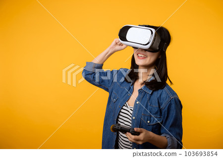 Young asian woman happy success wearing virtual reality headset game entertainment and holding joystick controller isolated on yellow background. Winner of cyber future video game concept. Young asian woman happy success wearing virtual reality headset game entertainment and holding joystick controller isolated on yellow background. Winner of cyber future video game concept. 103693854