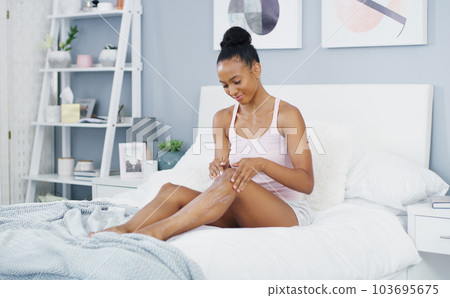 Woman, leg and moisturizer in bed for body care and cleaning in a home in morning lotion. Smile, relax and skincare with happy African female person with skin beauty and cosmetics cream in a house 103695675