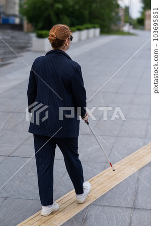 Blind businesswoman walking along tactile tiles with a cane.  103695851