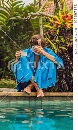 Sick man traveler. The man caught a cold on vacation, sits sad at the pool drinking tea and blows his nose into a napkin. His son is healthy and swimming in the pool. Travel insurance concept VERTICAL 103697388