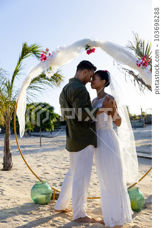 Side view of caucasian newlywed couple holding hands and standing at beach at wedding ceremony Side view of caucasian newlywed couple holding hands and standing at beach at wedding ceremony 103698288