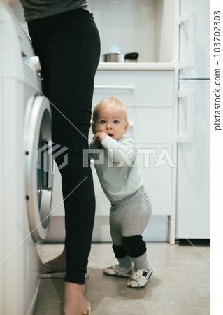 Little infant baby boy child hiding between mothers legs demanding her attention while she is multitasking, trying to do some household chores in kitchen at home. Mother on maternity leave Little infant baby boy child hiding between mothers legs demanding her attention while she is multitasking, trying to do some household chores in kitchen at home. Mother on maternity leave 103702303