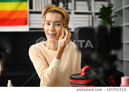 Pretty gay man beauty blogger applying base foundation on face with sponge, recording makeup tutorial video on smartphone 103703318