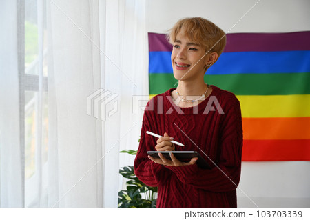 Positive teenage gay man in red sweater using digital tablet in bright living room with rainbow pride flag on background Positive teenage gay man in red sweater using digital tablet in bright living room with rainbow pride flag on background 103703339