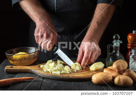 Before making the fries, the chef uses a knife to cut the raw potatoes into small pieces. Close-up of a cook hands while working in a restaurant kitchen 103707207