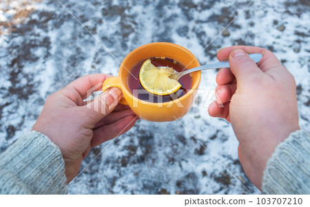 People hands hold an orange mug of hot herbal tea in cold winter. Prevention of viral diseases lemon tea. Spoon in person is hand. 103707210