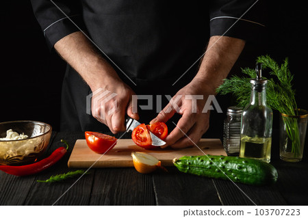 Chef hands with knife, cutting fresh red tomatoes for salad. Working environment in the restaurant kitchen. Fresh vegetables and oil on table Chef hands with knife, cutting fresh red tomatoes for salad. Working environment in the restaurant kitchen. Fresh vegetables and oil on table 103707223