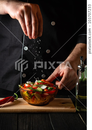 The cook sprinkles salted fresh vegetable salad in plate on a wooden table. Cooking healthy food in the kitchen in a restaurant 103707233