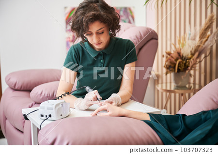 Manicurist removing cuticle on client hand with electric nail drill. Female manicure master sitting at the table while doing hardware manicure on woman hand. 103707323