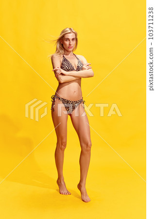 Beautiful young woman with perfect, slim, tanned body in stylish swimsuit against yellow studio background 103711238
