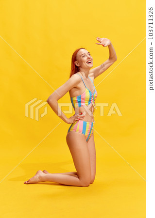 Beautiful, smiling redhead girl with slim, perfect body, wearing striped swimsuit, posing against yellow studio background 103711326