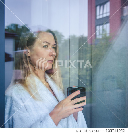 Woman in elegant robe drinking coffee in hotel room and standing near window. window reflection 103711952