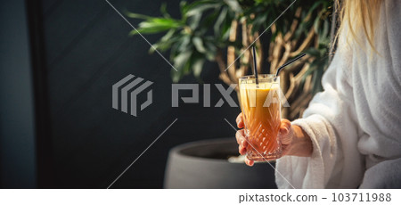 Woman in white bathrobe lying on sofa and relaxing with orange cocktail at home. Woman in white bathrobe lying on sofa and relaxing with orange cocktail at home. 103711988
