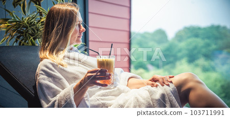 Woman in white bathrobe lying on sofa and relaxing with orange cocktail at home. Woman in white bathrobe lying on sofa and relaxing with orange cocktail at home. 103711991