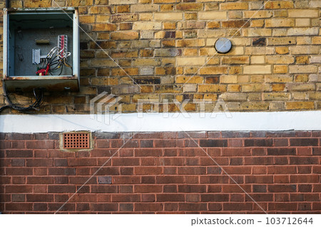 Bricks wall, with electricity / fuse box on it, opened, cover missing, cables visible. Bricks wall, with electricity / fuse box on it, opened, cover missing, cables visible. 103712644