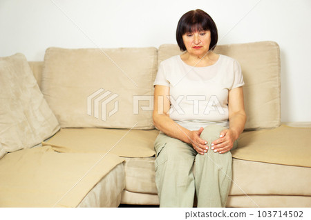 Unhappy senior woman suffering from knee pain on a sofa. People, health care and problem concept. Unhappy senior woman suffering from knee pain on a sofa. People, health care and problem concept. 103714502