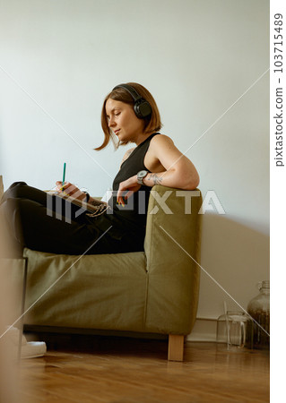 Female student listening to online lecture in headphones and making notes while studying from home 103715489