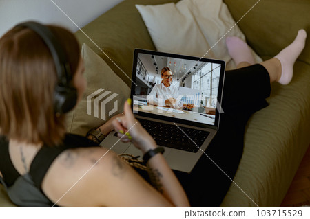 Woman in headphones sit on couch in living room and talking by video conference call with colleague Woman in headphones sit on couch in living room and talking by video conference call with colleague 103715529