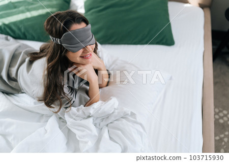 High-angle view of smiling young woman wearing sleeping mask lying in comfortable bed in luxury bedroom in hotel. Portrait of beautiful brunette female in pajama waking up enjoy good morning at home. 103715930