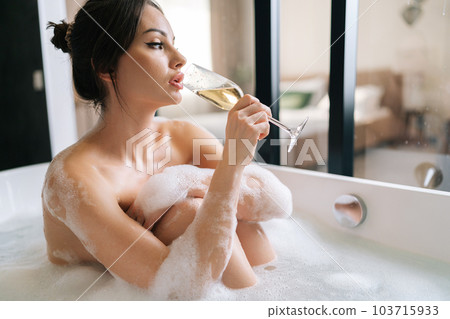 Side view of beautiful naked young woman drinking champagne sitting in foam bath enjoying alcoholic drink during luxurious recreation for weekend. Sexual brunette female relaxing in bathroom at hotel. 103715933