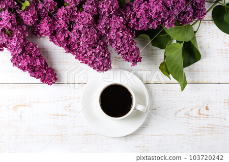 Cup of fresh black coffee and branch of blooming purple lilac with green leaves on white wooden background. Coffee break. Spring flowers. Flat lay, top view, copy space. 103720242